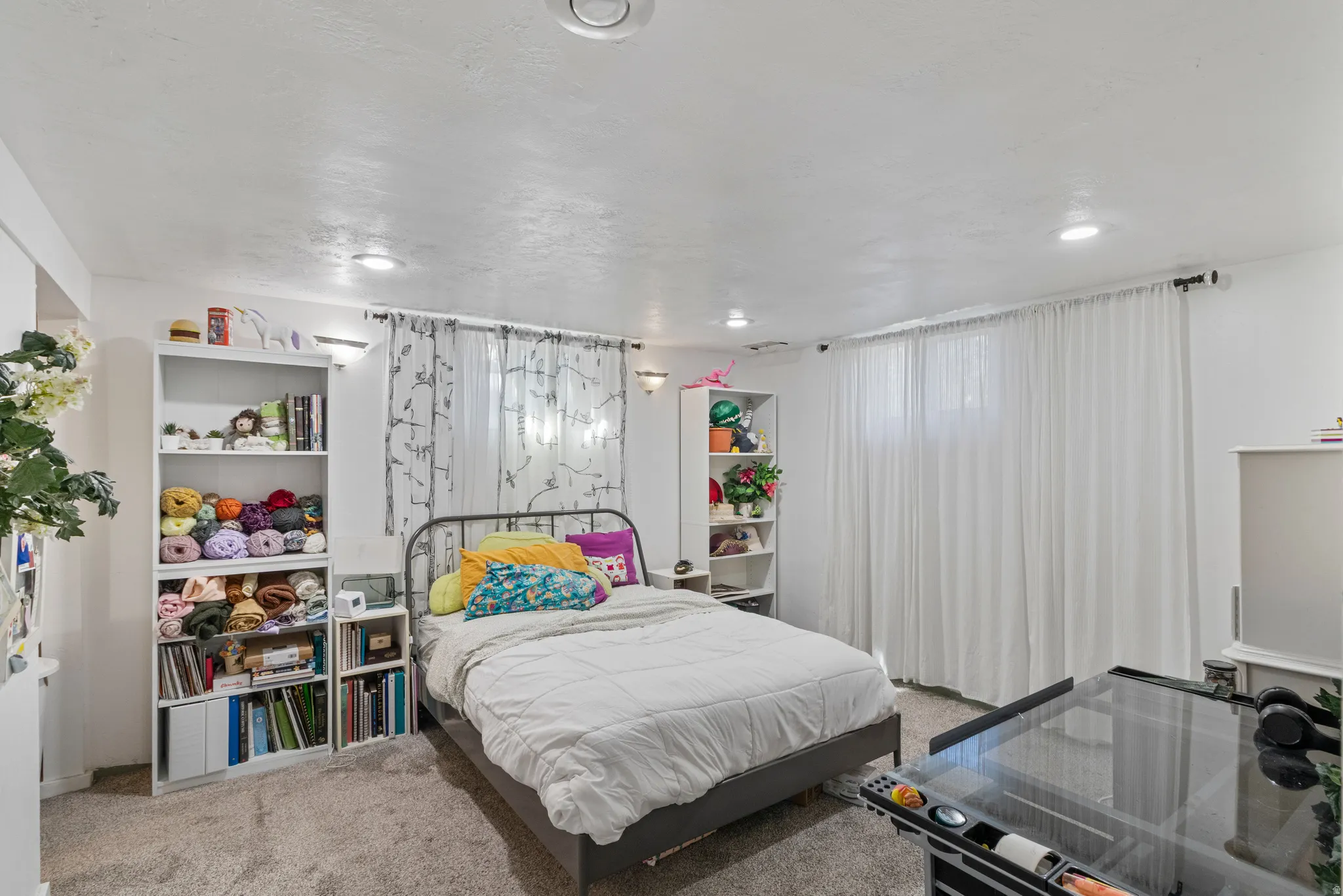 Spacious bedroom featuring recessed lighting and wall sconces, carpet flooring, a large window with sheer white curtains, and multiple built-in shelving units