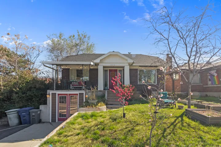 Brick facade residence featuring a covered front porch with iron railings, a bright red entry door, a sloped front lawn, and visible raised garden beds