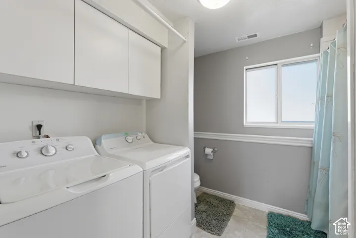 Dedicated laundry room featuring side-by-side washer and dryer appliances, ample overhead cabinetry for storage, large window with natural light, tile flooring, and a full bathroom with shower enclosure and vanity