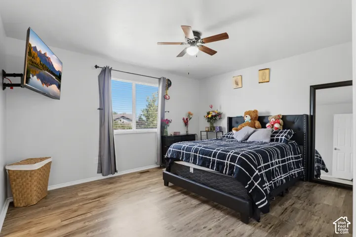 Spacious primary bedroom featuring a black upholstered bed frame, wood-finish flooring throughout, ceiling fan with integrated lighting, large window with thermal-insulated glass and gray curtain panels, and ample natural light