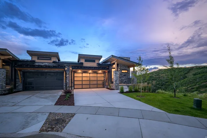 Contemporary architectural design with stone and wood siding, multiple garage doors including a full-view glass door, and a concrete driveway