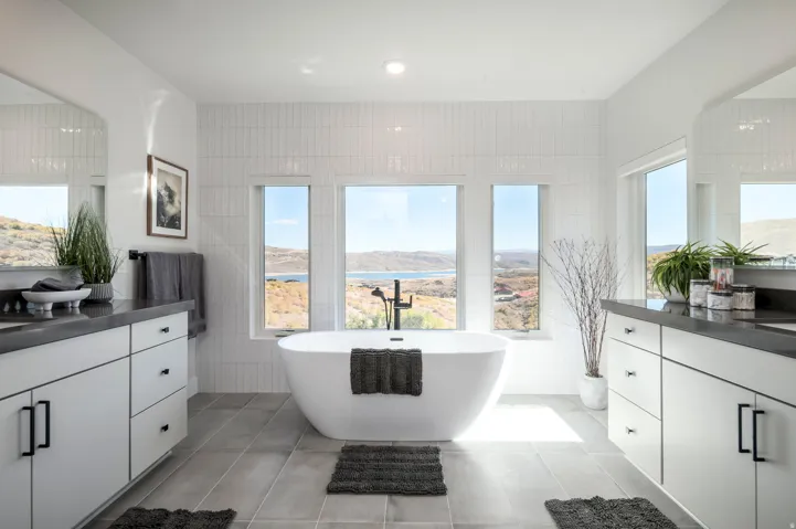 Spa-inspired primary bathroom featuring a freestanding soaking tub, dual vanities with white cabinetry and black countertops, large multi-panel windows framing expansive landscape vistas, tile flooring, and abundant natural light from recessed ceiling fix
