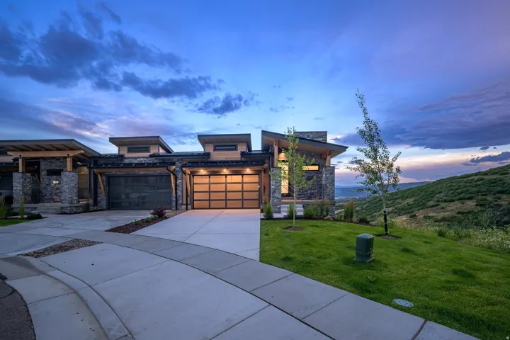 Modern architectural home featuring a three-car garage with gold-finish doors, wide concrete driveway with geometric scoring, mixed-material exterior combining stone and siding, contemporary roofline with multiple peaks, and manicured landscaping with mat