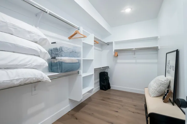 Spacious walk-in closet featuring custom white built-in shelving with multiple storage compartments, wooden hanging rods, warm wood-finish flooring, and abundant natural light from recessed ceiling fixtures