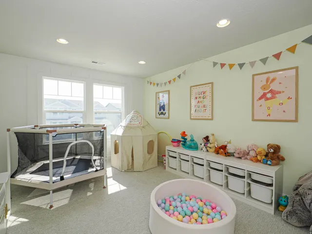 Bright, light-filled playroom featuring recessed ceiling lighting, large multi-pane windows with water views, soft neutral wall finishes, and built-in storage cabinetry with labeled bins