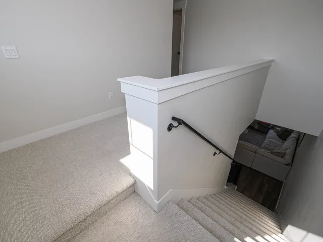 Carpeted staircase with a black metal handrail and white newel post, leading to a lower level with wood-finish flooring