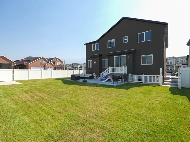 Modern two-story home with dark exterior siding and multiple windows, featuring a rear deck with white railings, expansive manicured lawn with mature grass, and white vinyl fencing
