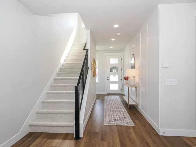 Wood-finish flooring, staircase with carpeted treads and black metal railing, vertical paneling detail on wall, recessed ceiling lighting, and a front door with frosted glass inserts