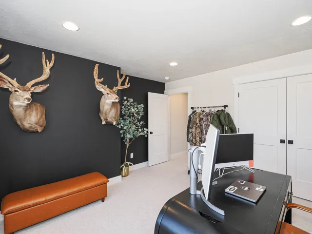 Sophisticated home office featuring recessed ceiling lighting, dark accent wall with decorative taxidermy mounts, carpeted flooring, built-in desk workspace with black surface, and white closet doors with hardware