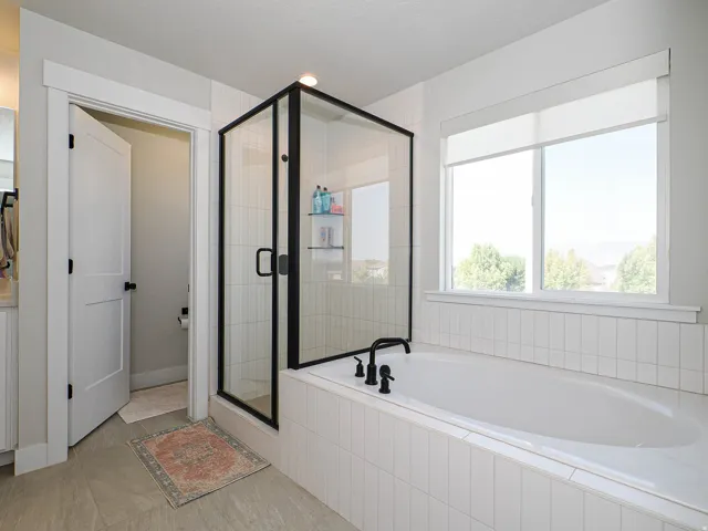 Spacious soaking tub with black fixtures, large window with roller shades, and a glass-enclosed shower with black trim