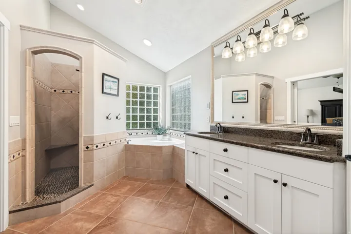 Spacious bathroom featuring a large soaking tub, walk-in shower with tiled bench, dual vanity with dark countertops, and a glass block window