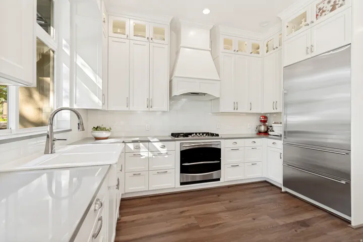 White cabinetry with glass-front uppers, stainless steel appliances, undermount sink with gooseneck faucet, light-toned countertops, and wood-finish flooring