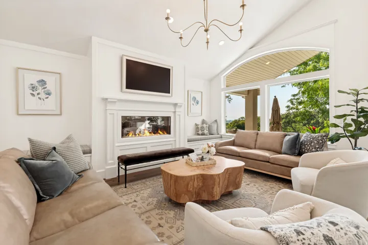 Vaulted ceiling with an elegant chandelier, a built-in linear fireplace with a detailed mantel, a window seat with storage, and large arched windows offering expansive outdoor views