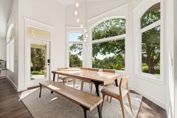 Bright dining area showcasing expansive transom and picture windows framing mature landscape views, light wood-finish dining table with bench seating, mid-century modern chairs, pendant lighting, dark wood-finish flooring, neutral area rug, and direct acc