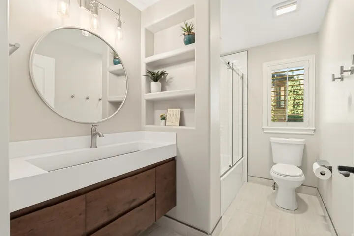 Modern primary bathroom featuring a circular framed mirror, floating wood-finish vanity with rectangular vessel sink, white quartz countertop, chrome faucet, built-in shelving with decorative accents, walk-in shower with glass enclosure, and light wood-fi