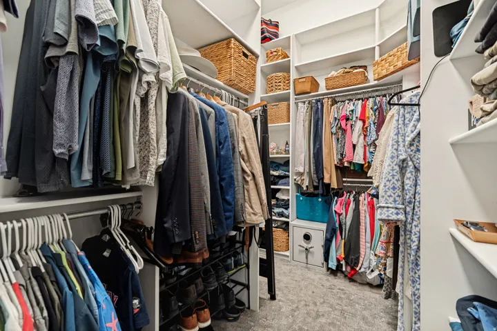 Spacious walk-in closet featuring dual clothing rods with multiple hanging sections, woven storage baskets on upper shelving, built-in white cabinetry with open shelving for folded garments, and neutral carpeted flooring throughout