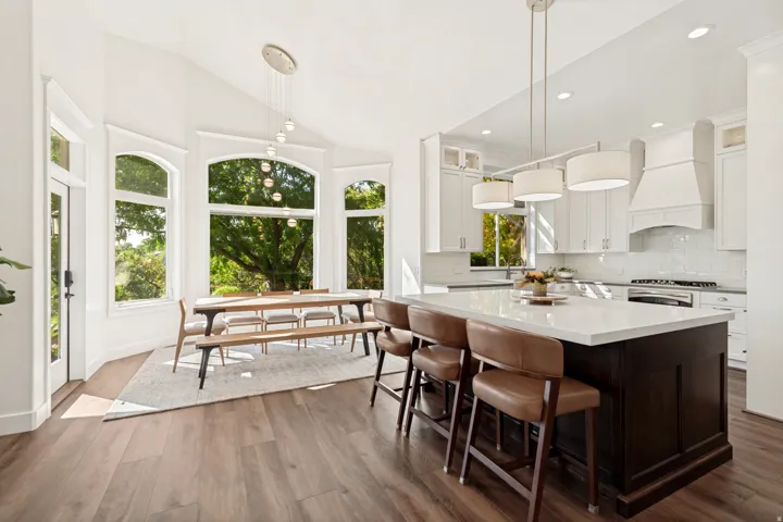Bright, light-filled kitchen and dining space featuring expansive multi-pane windows overlooking mature landscaping, wide-plank wood-finish flooring throughout, a dark-stained island with waterfall countertop and seating for multiple guests, white cabinet