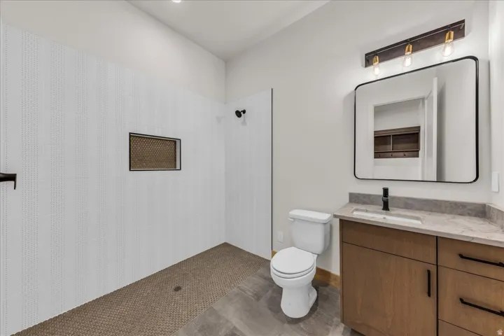 Walk-in shower with vertical white tile and built-in niche, wood-finish vanity with stone countertop, rectangular undermount sink, and matte black fixtures, framed mirror with integrated lighting, and mosaic tile shower flooring