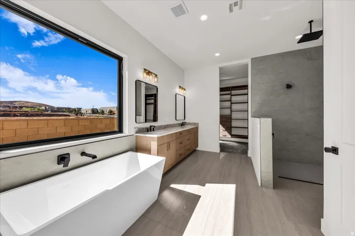 Freestanding soaking tub with wall-mounted matte black fixtures, dual vanity with wood-finish cabinetry and stone countertops, and an open shower with a dark-tiled floor