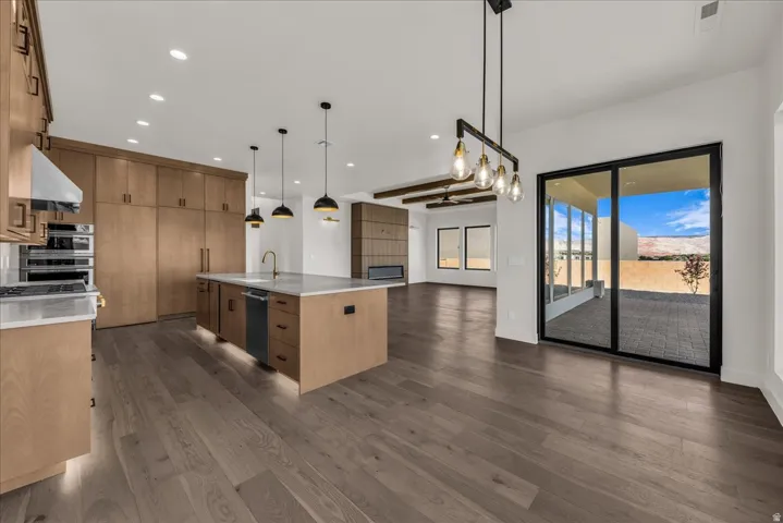 Open-concept kitchen and living area featuring natural wood cabinetry, a large center island with waterfall countertop, stainless steel appliances, wide-plank wood-finish flooring, pendant lighting, and expansive glass sliding doors opening to outdoor spa