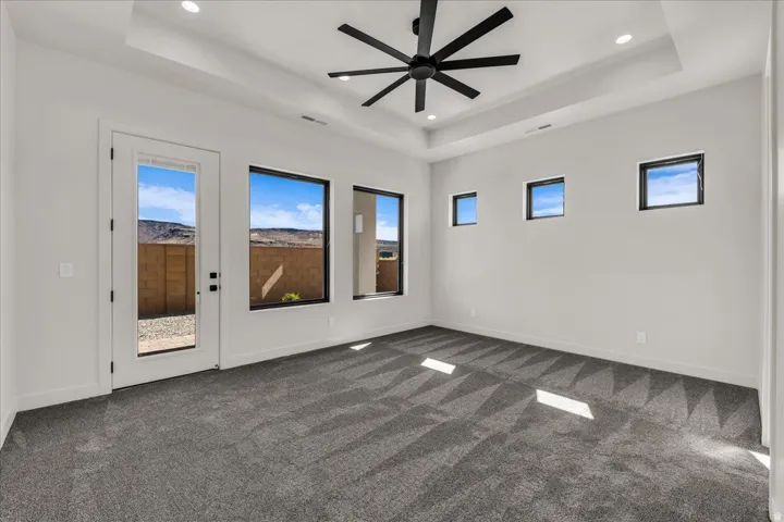 Tray ceiling with recessed lighting, modern ceiling fan, gray patterned carpeting, multiple windows, and a glass-paneled door