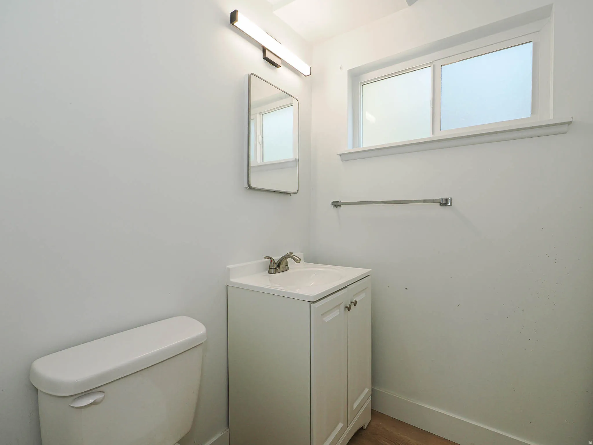 Clean and bright bathroom featuring a white porcelain toilet, pedestal-style vanity sink with chrome faucet, framed mirror, towel bar, and abundant natural light from dual windows