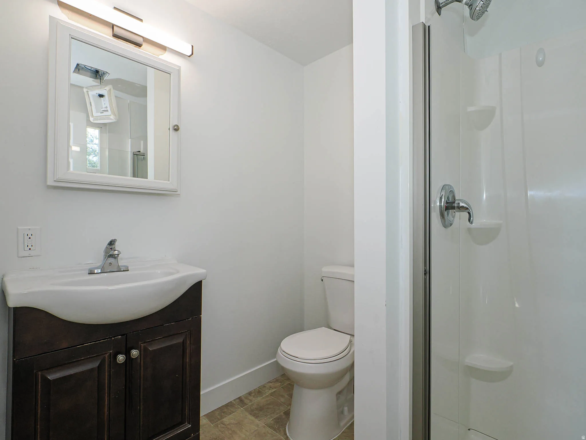 Bright bathroom featuring a dark wood vanity with curved basin sink, mirrored medicine cabinet with integrated lighting, ceramic toilet, and frameless glass shower enclosure with tile surround