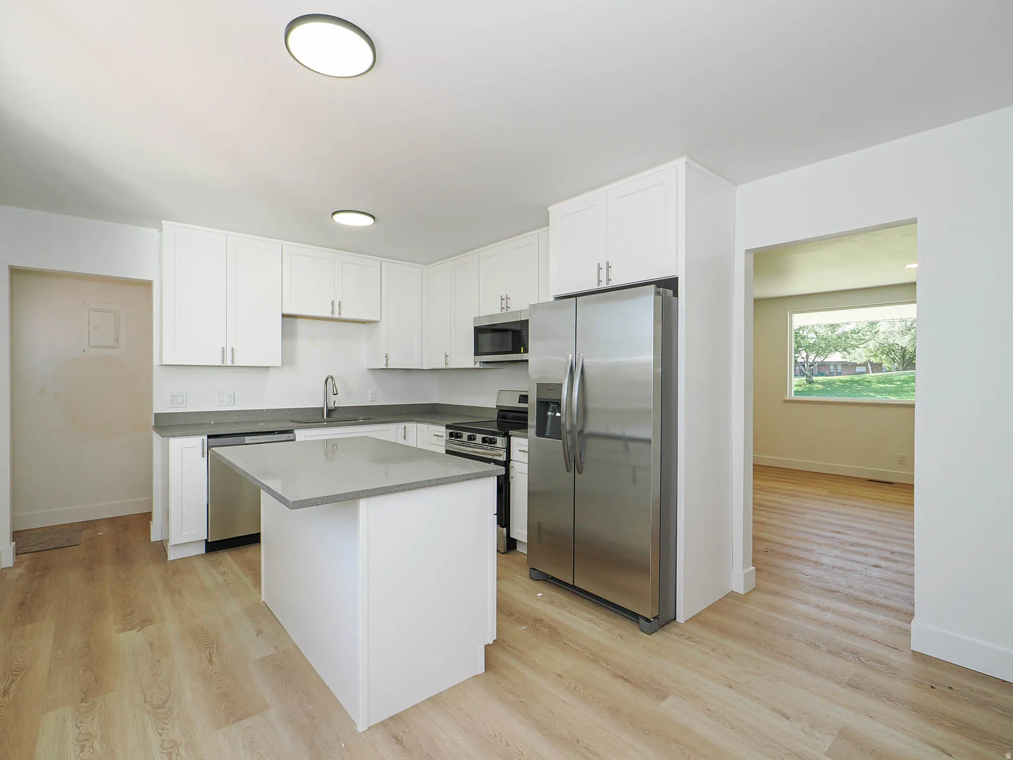 Contemporary kitchen with white shaker cabinetry, stainless steel appliances, light wood-finish flooring, a central island with a solid surface countertop, and recessed ceiling lighting