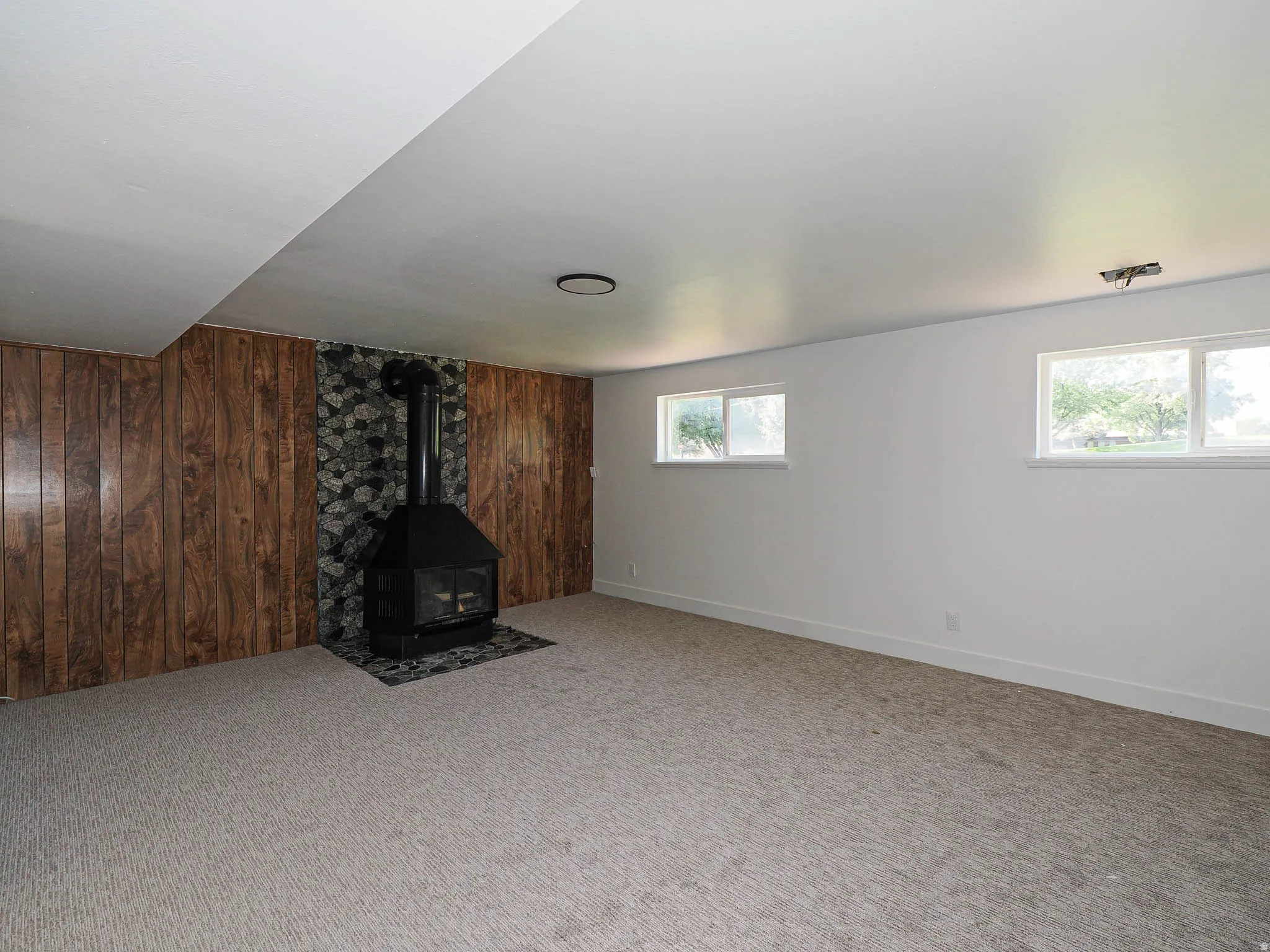 Carpeted room featuring a freestanding fireplace with a stone surround, wood-paneled accent wall, and multiple windows