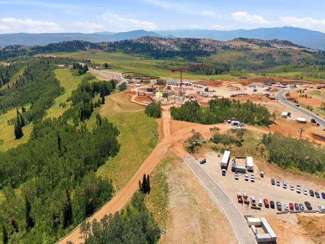 Expansive mountain landscape featuring a large development area with ongoing construction, surrounding mature evergreen trees, and a paved road system