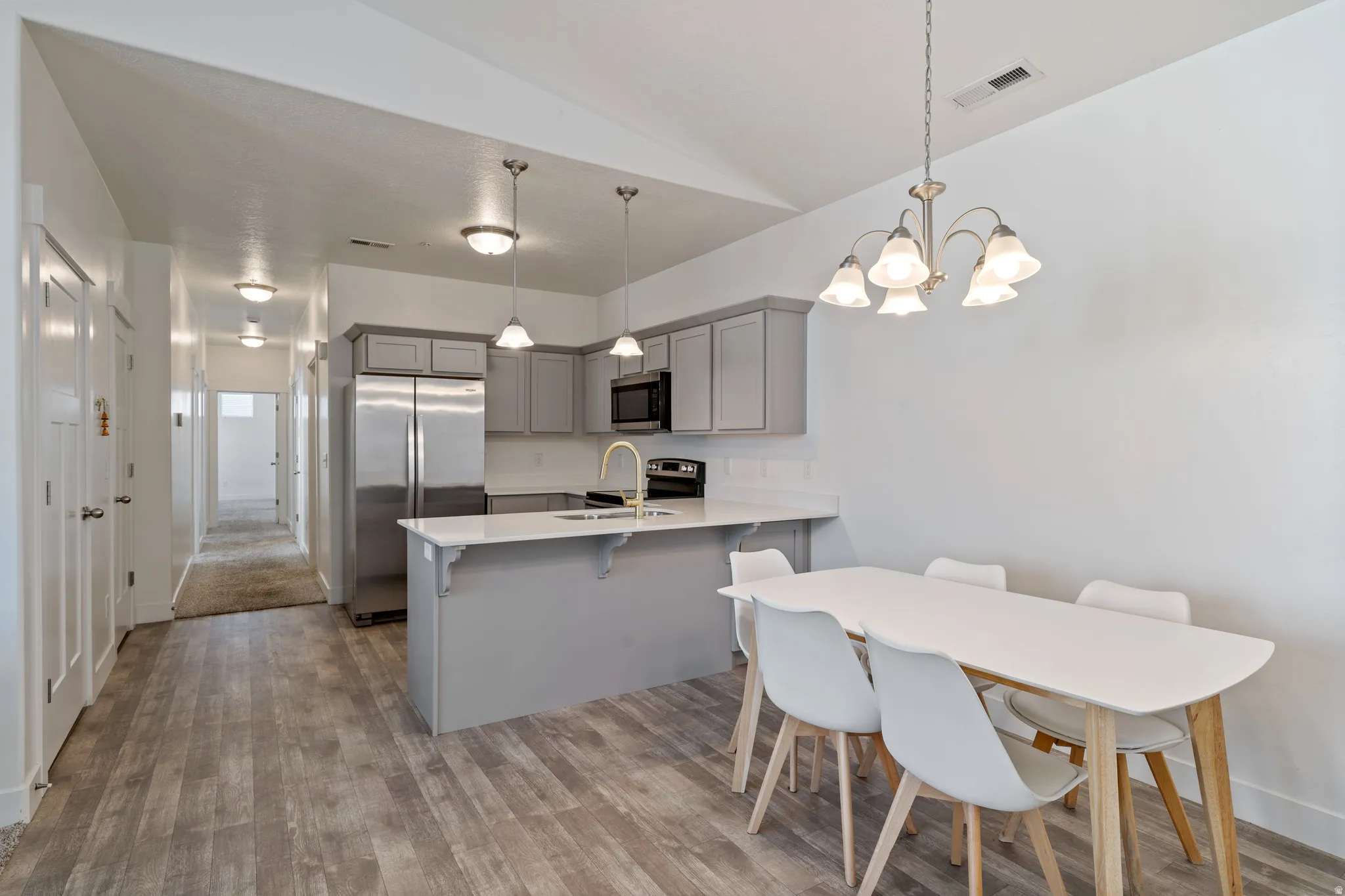 Open-concept kitchen and dining area featuring stainless steel appliances, gray cabinetry with soft-close doors, a quartz countertop island with seating, LVP flooring throughout, and a multi-light pendant chandelier suspended above the dining table