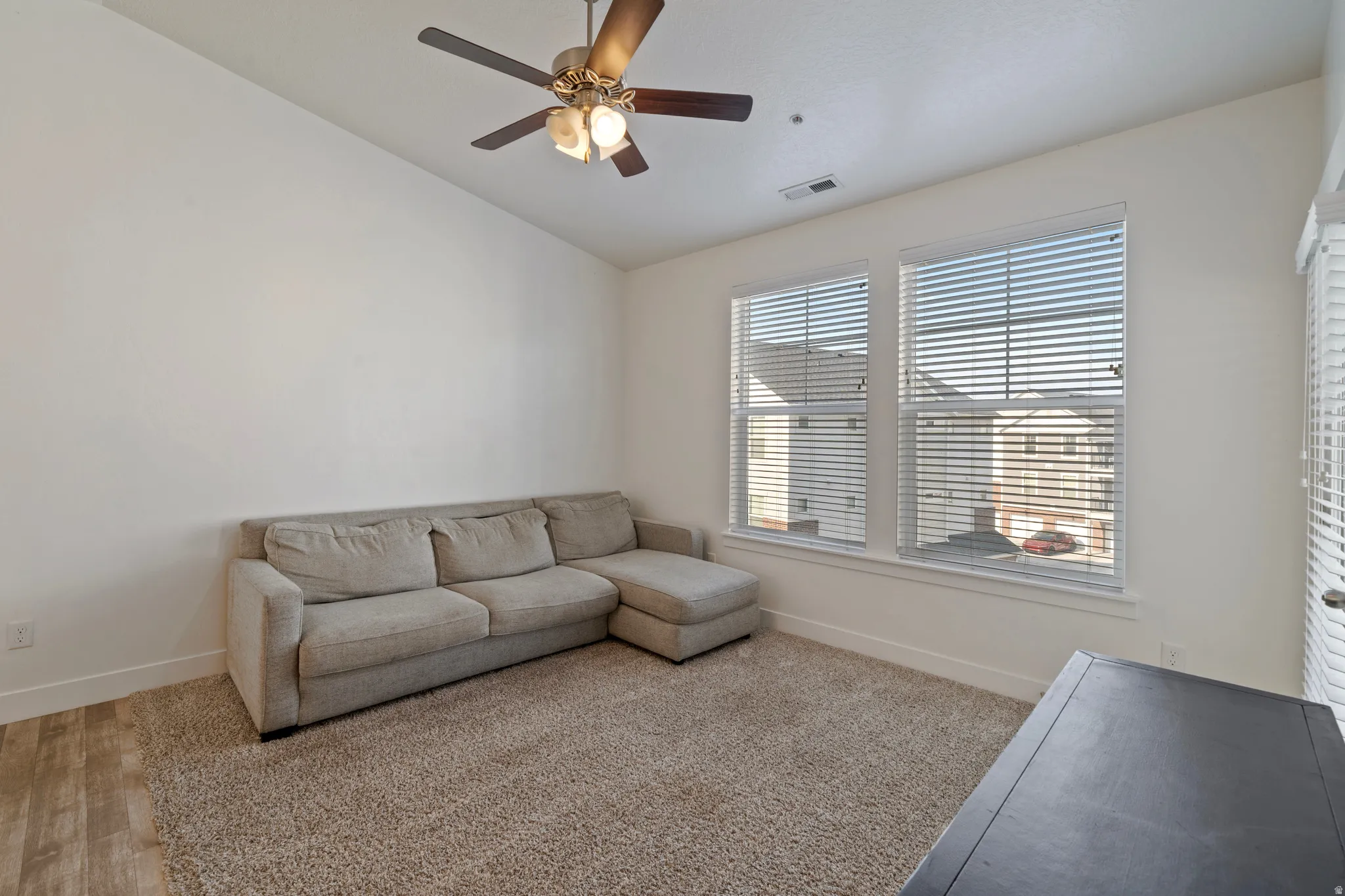 Bright, light-filled living room featuring expansive windows with plantation shutters, wood-finish flooring, neutral wall finishes, and a ceiling fan with integrated lighting