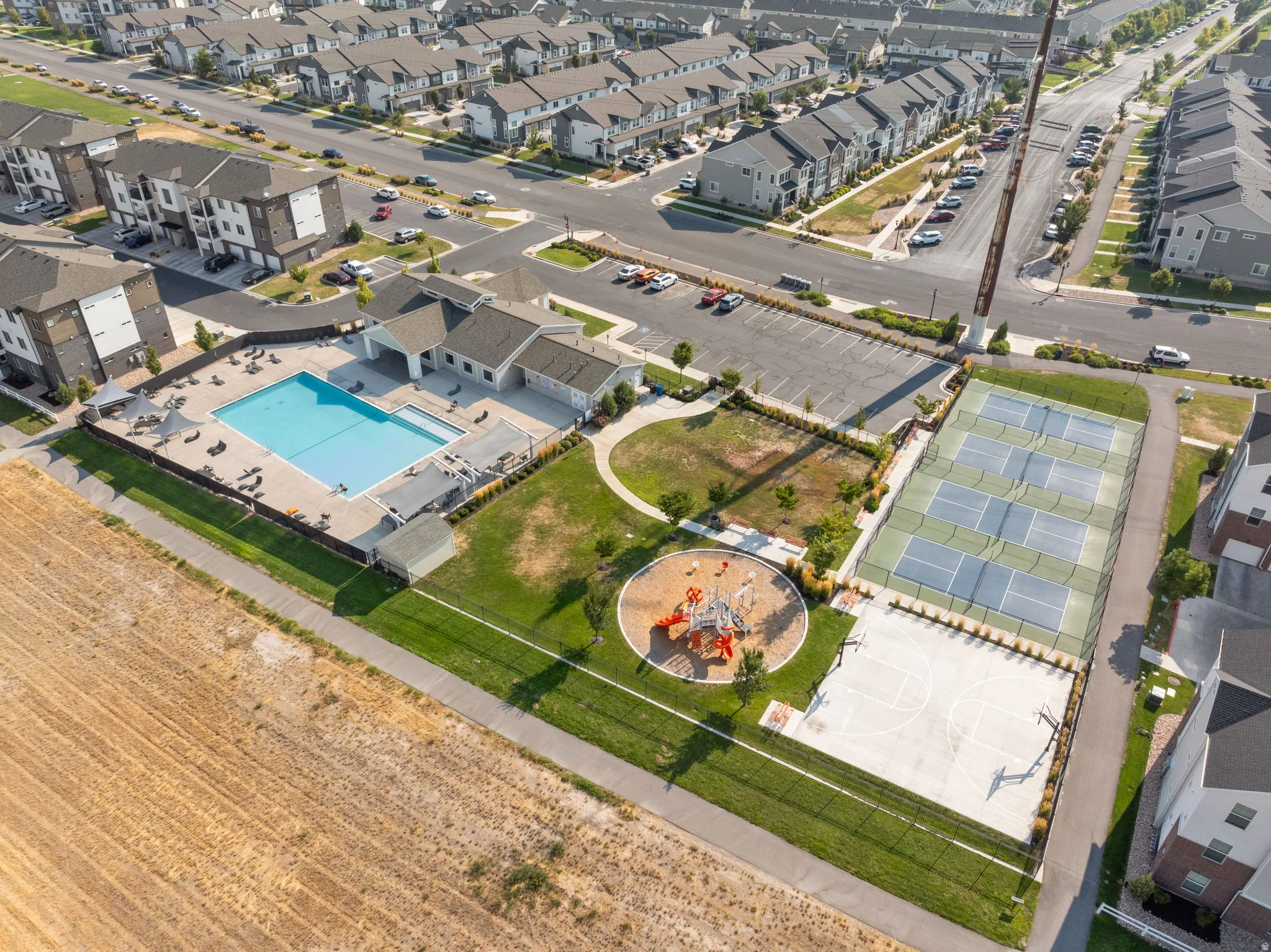Community amenities featuring a resort-style swimming pool with surrounding deck, lighted pickleball courts, a playground with recreational equipment, and manicured landscaping with mature trees bordering agricultural land