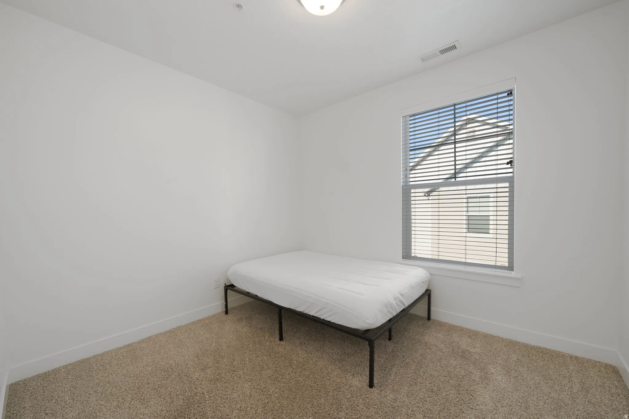 Bright thrid bedroom featuring neutral wall finishes, carpeted flooring, and a large single-hung window with horizontal blinds providing natural light and ventilation