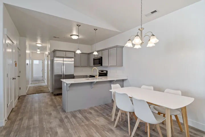 Open-concept kitchen and dining area featuring stainless steel appliances, gray cabinetry with soft-close doors, a quartz countertop island with seating, LVP flooring throughout, and a multi-light pendant chandelier suspended above the dining table