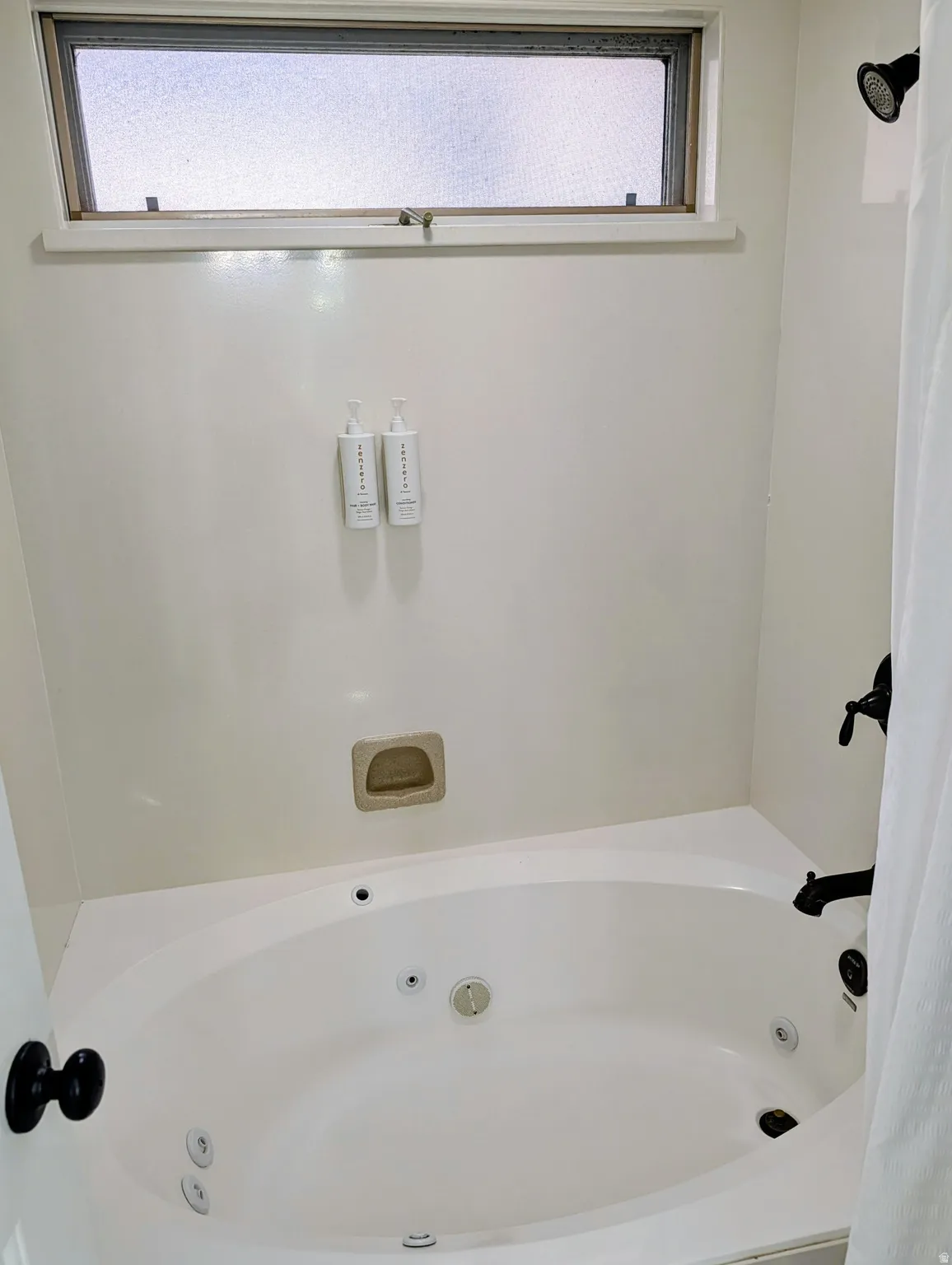 Spa-inspired primary bath featuring a curved whirlpool tub with integrated jets, transom window with natural light, dual wall-mounted dispensers, recessed shelving niche, and crisp white tile surround with dark hardware accents