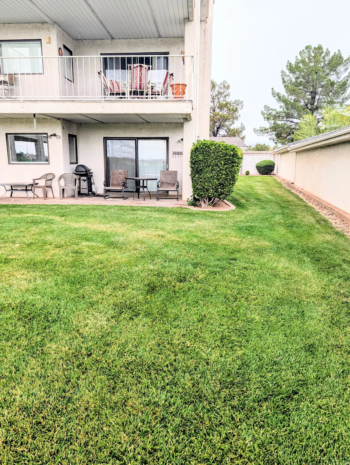 Expansive manicured lawn with mature landscaping and privacy fencing, complemented by a two-level residence featuring a ground-level patio area with sliding glass doors, upper-level balcony with metal railing, and established greenery throughout the groun