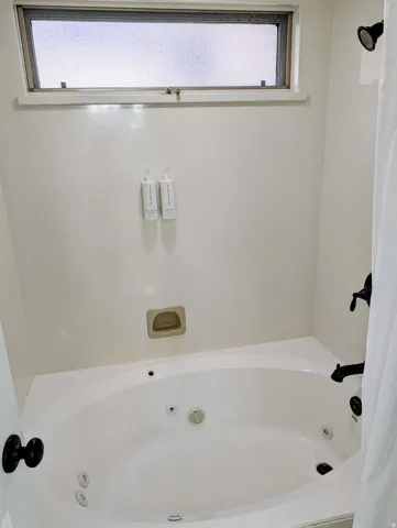 Spa-inspired primary bath featuring a curved whirlpool tub with integrated jets, transom window with natural light, dual wall-mounted dispensers, recessed shelving niche, and crisp white tile surround with dark hardware accents