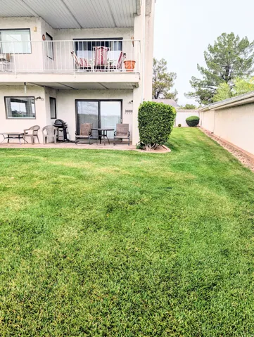 Expansive manicured lawn with mature landscaping and privacy fencing, complemented by a two-level residence featuring a ground-level patio area with sliding glass doors, upper-level balcony with metal railing, and established greenery throughout the groun