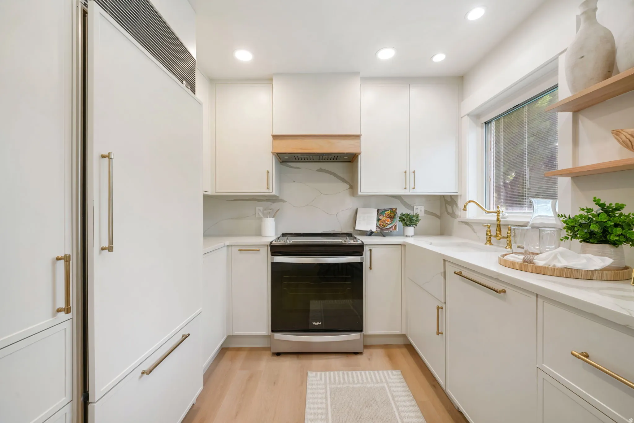 White cabinetry with brass hardware, undermount sink with brass faucet, light wood-finish flooring, white stone countertops, and built-in stainless steel range