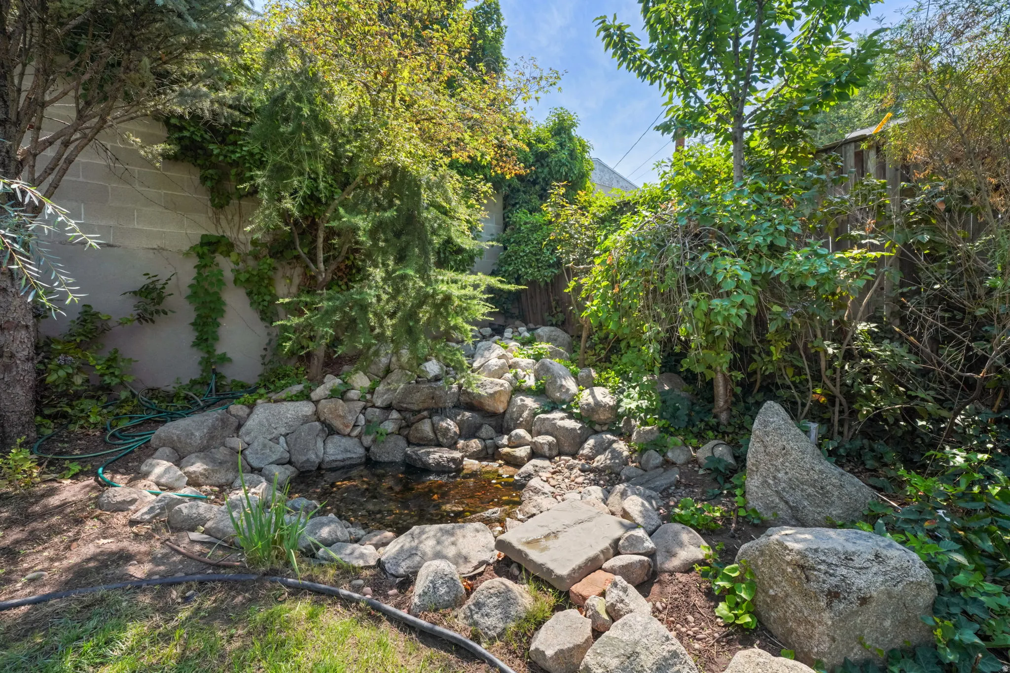 Stone-lined water feature with established landscaping, mature trees, and a block retaining wall