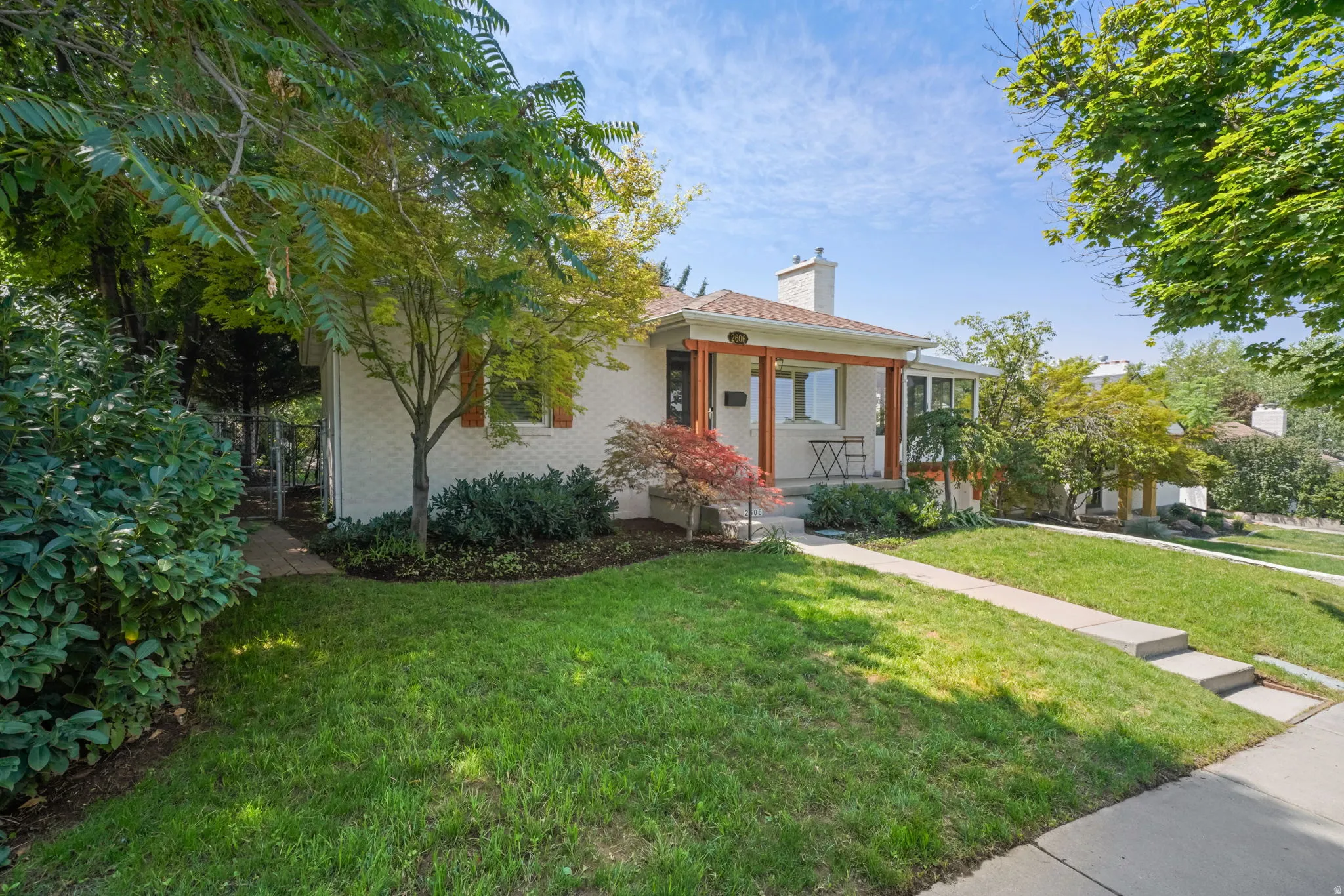 White brick exterior with a covered porch, mature trees, concrete walkway, and landscaped garden beds