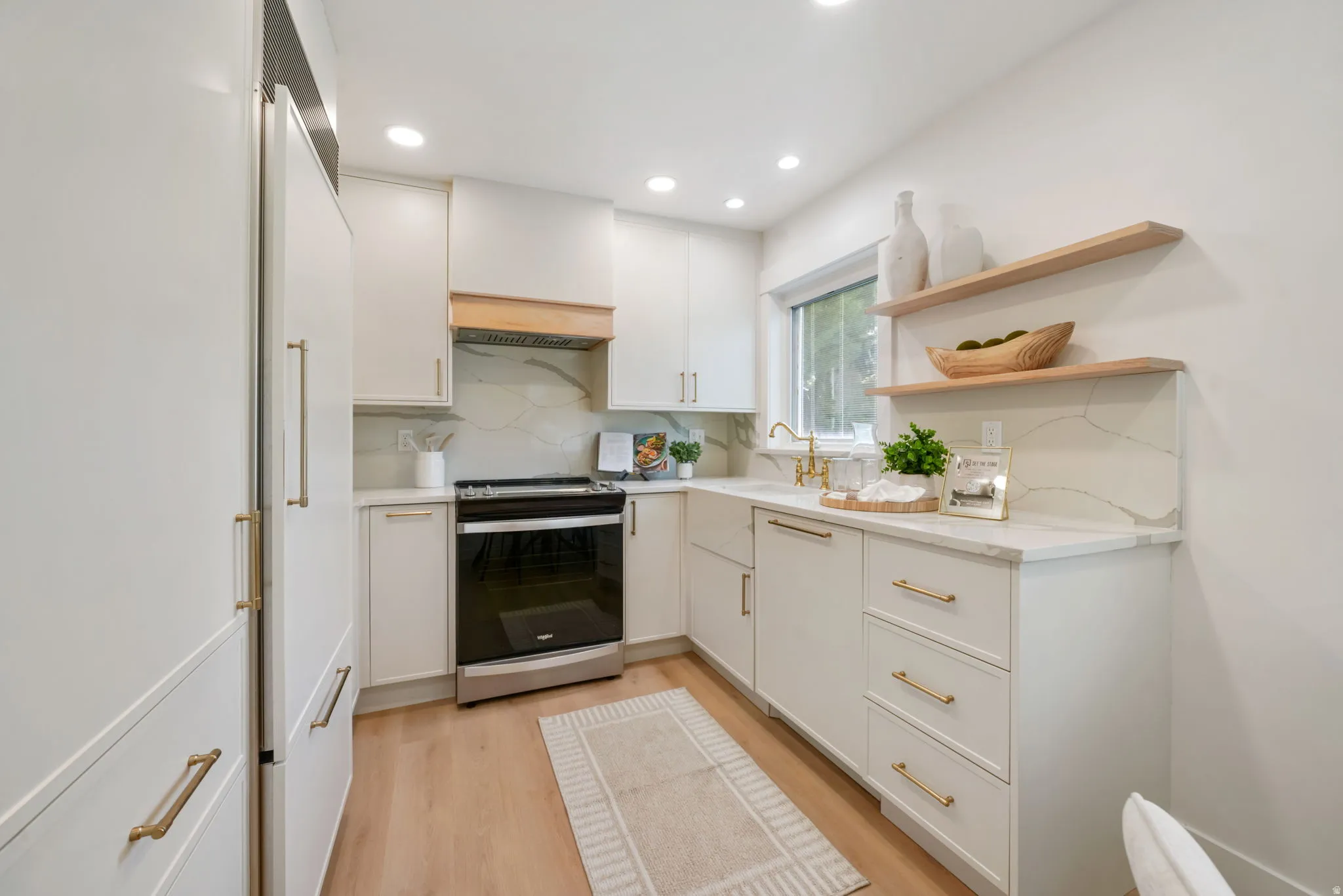 Recessed lighting illuminates the kitchen featuring wood-finish flooring, white cabinetry with brass hardware, a white range, and a white countertop with a matching backsplash