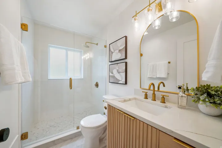 Glass-enclosed shower with a rainfall showerhead, a single vanity with a light wood-finish cabinet, a white stone countertop, and gold-finish fixtures