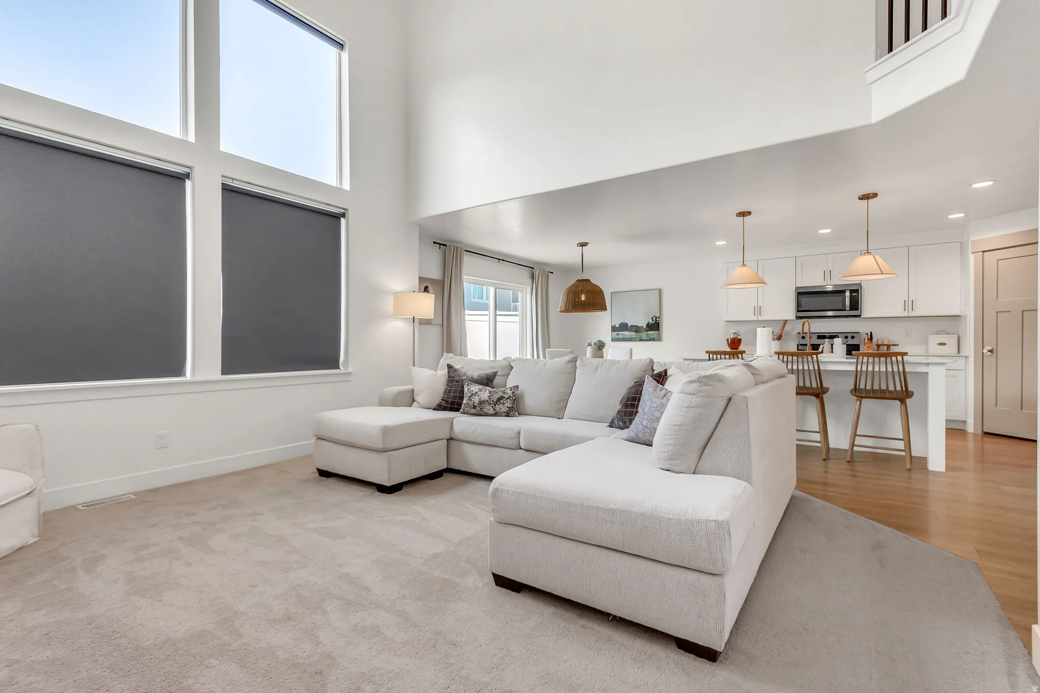 Two-story living area with high ceilings and large windows, open to a kitchen featuring white cabinetry, light wood-finish flooring, and pendant lighting