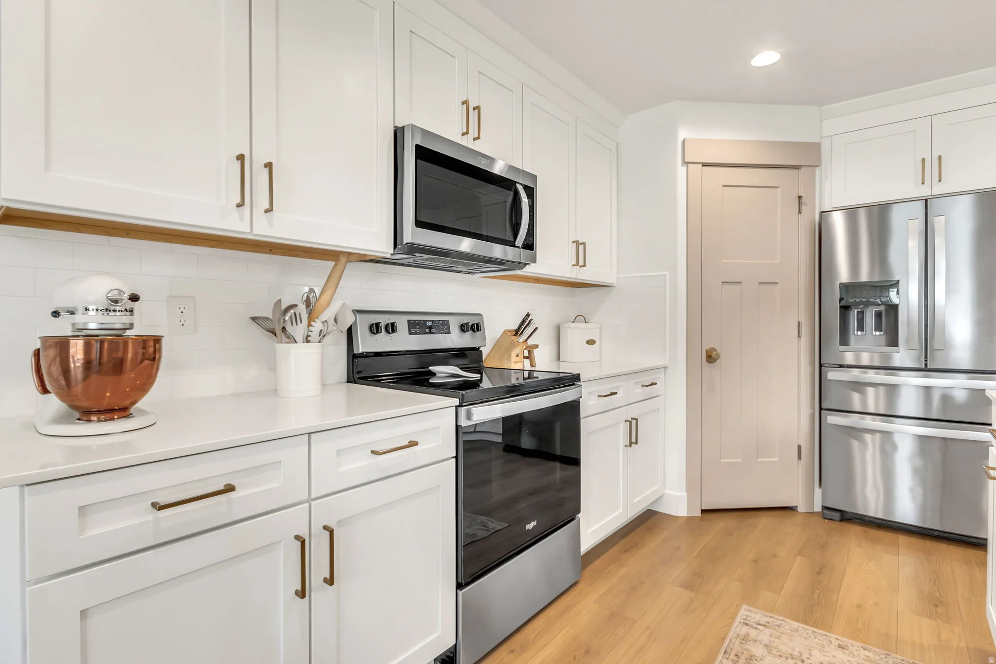 Kitchen featuring white shaker-style cabinetry, stainless steel appliances, a white subway tile backsplash, light-toned wood-finish flooring, and solid surface countertops