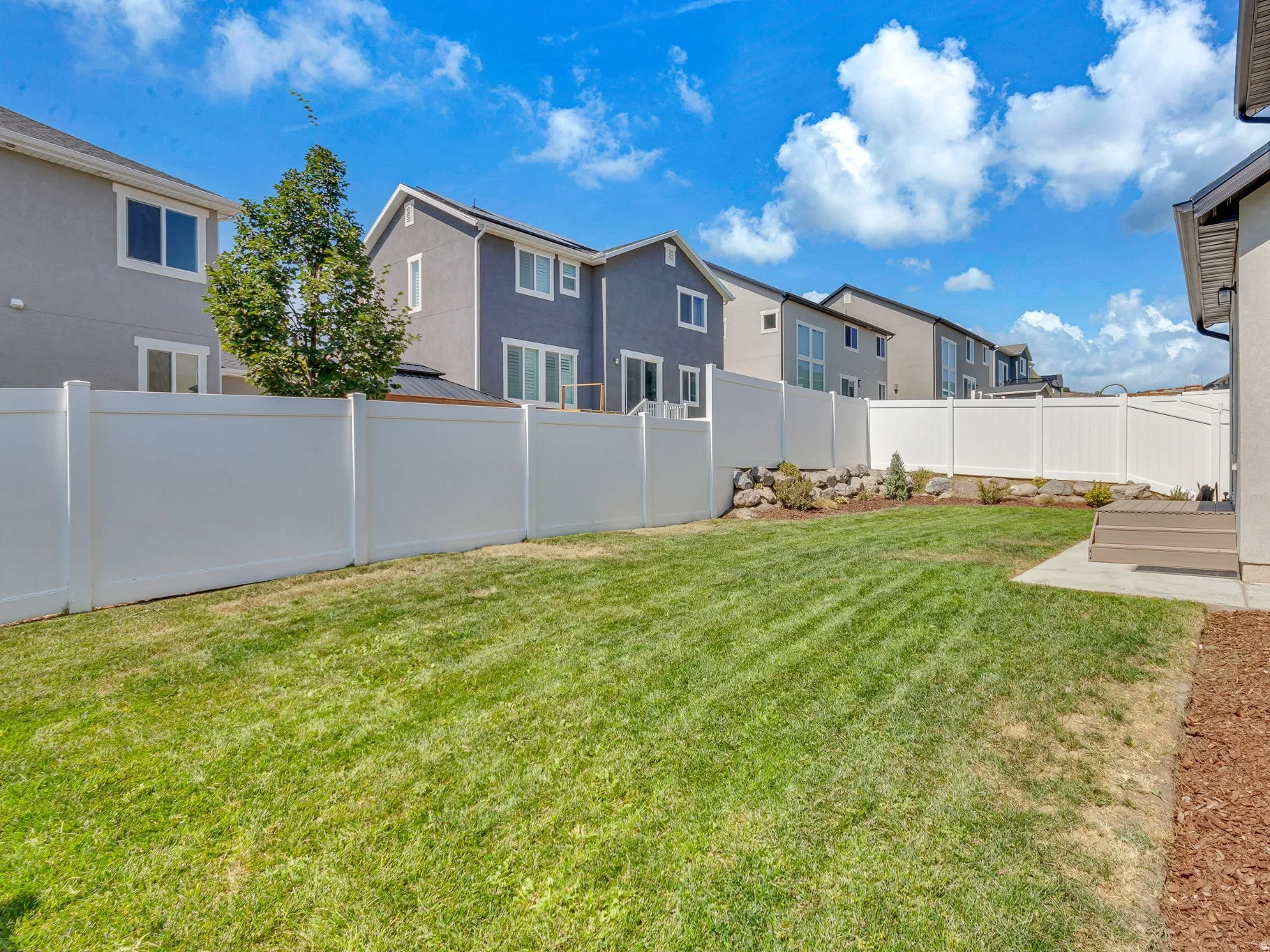 Expansive backyard featuring a manicured lawn, white privacy fencing, a concrete patio, and a decorative rock and shrub border