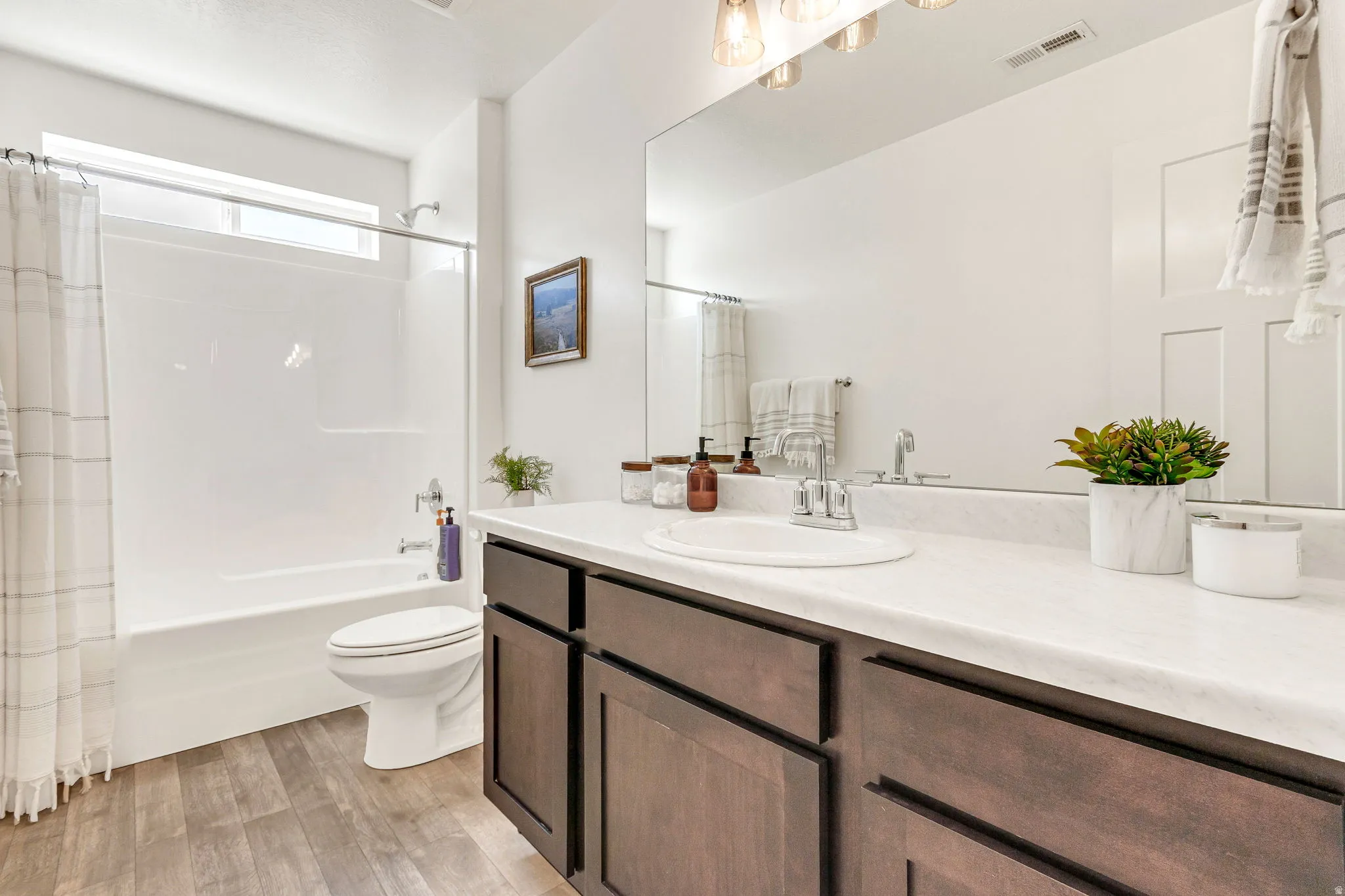Bathroom featuring a wood-finish flooring, a white vanity with dark wood-finish cabinetry, a large vanity mirror, and a full bathtub with an overhead window
