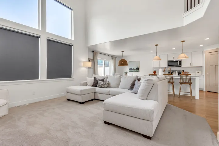 Two-story living area with high ceilings and large windows, open to a kitchen featuring white cabinetry, light wood-finish flooring, and pendant lighting