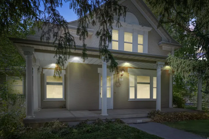 Classic facade featuring a covered porch with decorative columns, brick-finish siding, a gabled roofline with shingle accents, and illuminated windows